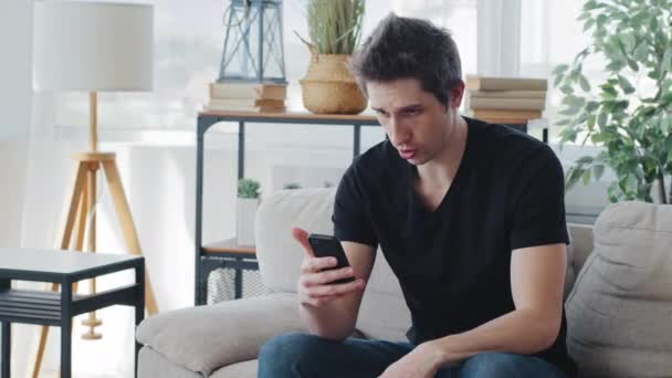 Enojado molesto caucásico adulto hombre emocional millennial chico usa camiseta negra sentado en el sofá hace videollamada conferencia en línea utilizando el teléfono móvil habla gritos en concepto de conflicto de cámara de teléfono inteligente — Vídeo de stock