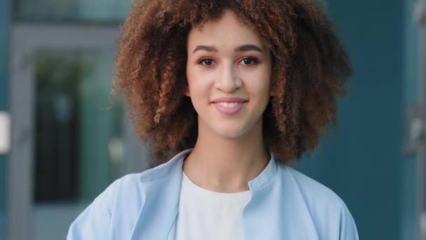 Female portrait beautiful young ethnic afro american girl mixed race woman lady model student with curly hair african hairstyle standing outdoors smiling toothy looking at camera waving hello greeting — Stock Video