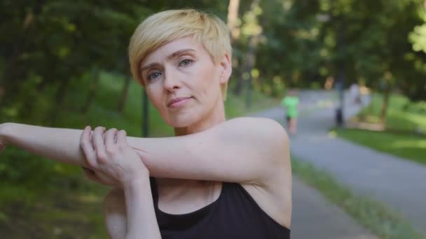 Portret van een mooie vrouw van middelbare leeftijd blond volwassen vrouw sterke vrouwelijke atleet trainer doen sport werk training strekt spieren van armen stretching oefening buiten staan in het park — Stockvideo