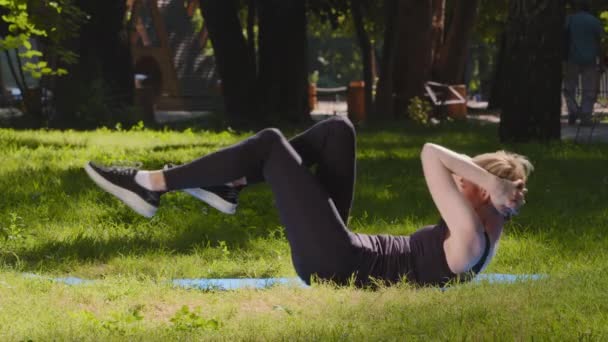 アスレチックスレンダーな強い白人女性トレーナー女性の女性は公園で屋外の芝生の上にマットの上に横たわる足や体を曲げ腹筋トレーニングのための物理的な演習を行う力 — ストック動画