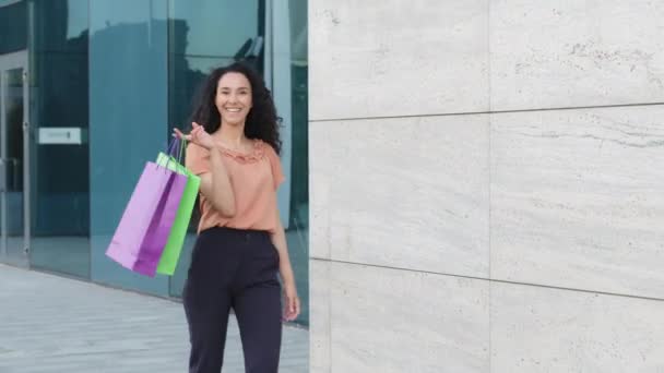 Молодая красивая улыбающаяся счастливая латиноамериканка покупательница потребительница женщина, идущая по городской улице возле здания торгового центра несет бумажные пакеты с покупками новых товаров одежды кружит радуется шоппинг — стоковое видео