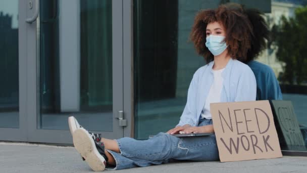 Lockige Geschäftsfrau Afroamerikanerin professionelle Spezialistin entlassen Arbeiter mit Gesichtsschutz medizinische Maske sitzen auf der Straße im Freien mit Zeichen Notwendigkeit Arbeit. Coronavirus-Pandemie bei Arbeitslosigkeit — Stockvideo