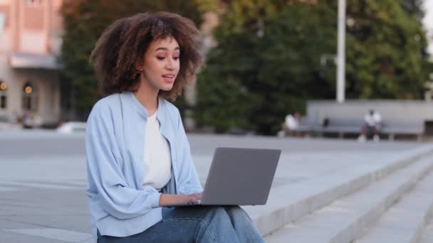 Afro-americana com cabelo encaracolado senhora africana menina estudante freelancer sentado ao ar livre na cidade usando laptop on-line vídeo conferência chat acenando Olá adeus falando com webcam — Vídeo de Stock