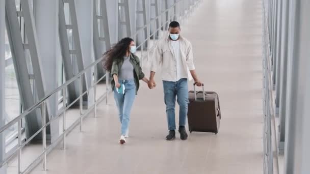 Счастливые счастливые многорасовые пары африканский мужчина и латиноамериканка в защитных медицинских масках на лицах бегущие танцы прыжки от радости веселья путешествия отпуск нести чемодан взявшись за руки в аэропорту — стоковое видео