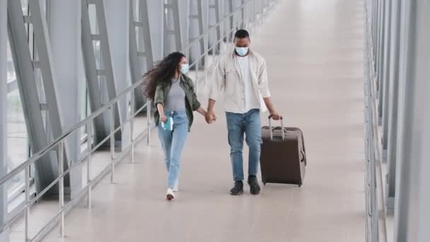 Feliz pareja multirracial alegre hombre africano y mujer hispana con máscaras médicas protectoras en las caras corriendo bailando saltando de alegría divertidas vacaciones de viaje llevar maleta de la mano en el aeropuerto — Vídeos de Stock