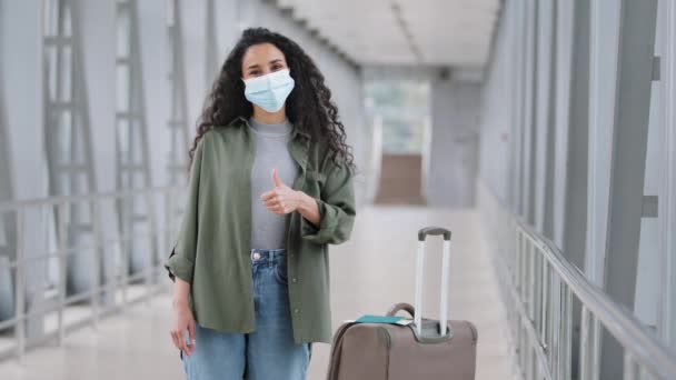 Латиноамериканская девушка-путешественница носит защитную маску для лица стоит на терминале аэропорта с чемоданом, глядя в камеру показывает, как большие пальцы вверх поддерживает рекомендует безопасное путешествие в пандемии — стоковое видео