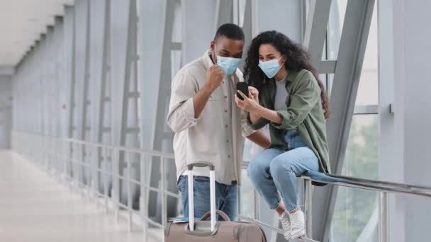 Pareja multiracial mujer hispana y hombre afroamericano con máscaras médicas sentados en el aeropuerto con maleta mirando el teléfono móvil ganar éxito haciendo gesto de victoria con la mano feliz con el vuelo — Vídeos de Stock