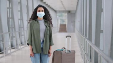 Kıvırcık saçlı İspanyol kadın, kadın yüzüne koruyucu maske takıyor havaalanı tren istasyonunda bavulun yanında pandemik karantinada seyahat eden kameralara bakıyor.