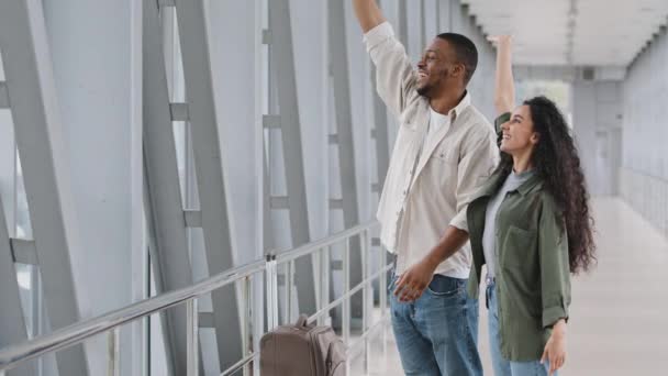 Casal multirracial mulher hispânica e homem africano viajantes com mala encontrar amigos no aeroporto após avião chega ao terminal à espera de embarque olhando para a janela acenando mãos Olá com alegria — Vídeo de Stock