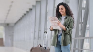 Yalnız, mutlu, genç, İspanyol, aşılanmış güzel, kadın turist, havaalanı terminalinde elinde bavuluyla karantinanın bitiş yolunu bulmak için haritaya bakıyor.