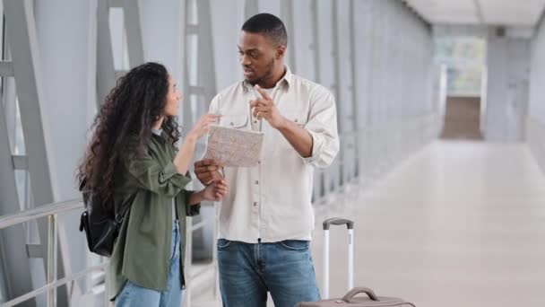 多人種間の若いカップルの観光客ヒスパニック系の女性の妻とアフリカ人男性アフロ夫は、地図検索方向道路を見て新国に到着した後、スーツケースと空港ターミナルに立つ主張 — ストック動画