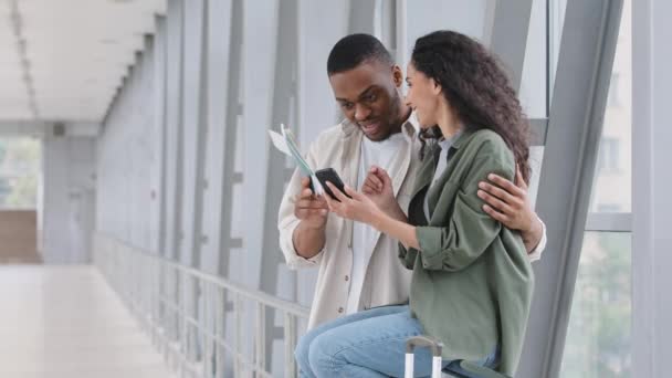 Multiracial par afrikansk man och latinamerikansk kvinna med flygbiljetter pass sitter i flygplatsterminalen med navigator app mobiltelefon titta på foto från resa surfa nätverk med smartphone — Stockvideo