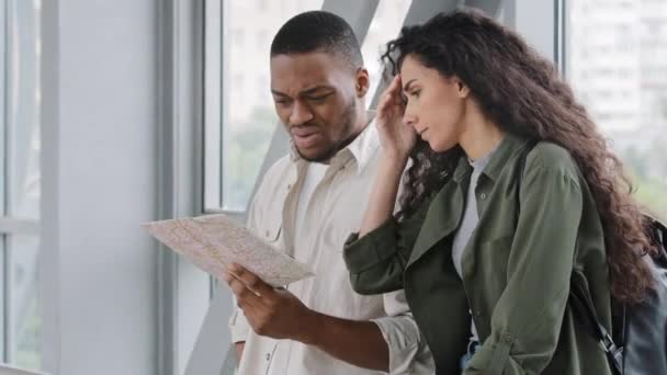 Upset sad multiracial couple African man and Hispanic woman lost in new country stand at airport looking at map searching for way direction worried. Afro guy tourist asks girl for help with route — Vídeo de stock