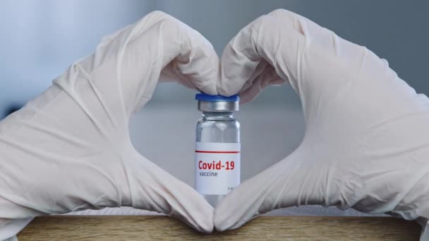 Primer plano de la mano femenina en guantes médicos de látex blanco que hacen que la forma del corazón muestre signos de amor esperanza confianza aprobación cerca del frasco de dosis de vacuna de covid coronavirus pandemia remedio cura vial medicamento — Vídeo de stock