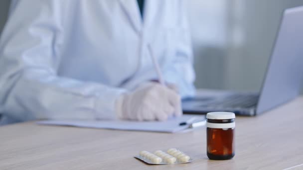 Крупним планом пляшка медичних таблеток стоїть на дерев'яному столі в клінічній лікарні на тлі розмитого неорієнтованого лікаря медсестри медичного працівника в уніформі письмового призначення, що працює на ноутбуці — стокове відео
