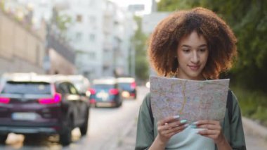 Mutlu gülümseyen Afro-Amerikan kadın turist elinde kağıt haritayla güneşli yaz gününde. Genç melez kadın seyahat blogcusu şehir merkezinde gezmek için rehber kitap kullanıyor.
