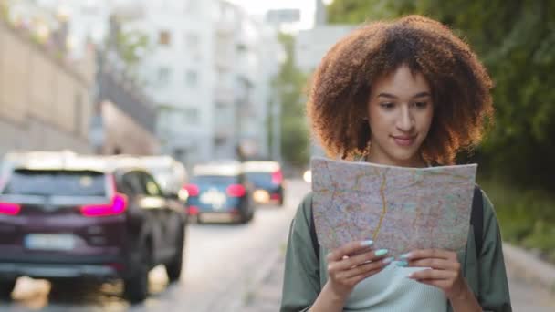 Feliz sorrindo atraente afro-americano mulher turista com papel mapa em mãos no verão dia ensolarado. Jovem mista blogueira de viagem feminina viajando no centro da cidade usa guia para passear — Vídeo de Stock