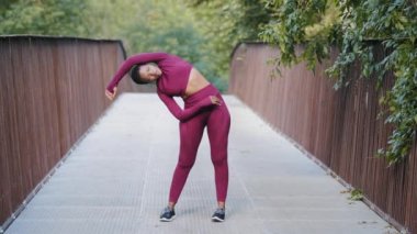 Spor giyim sektöründe tek başına egzersiz yapan atletik genç bir kadın. Afrikalı siyahi yetişkin kadın parkta sabah koşusu yaparken esneme egzersizleri yapıyor. Pilates, kardiyo eğitimi, yoga ve fitness konsepti
