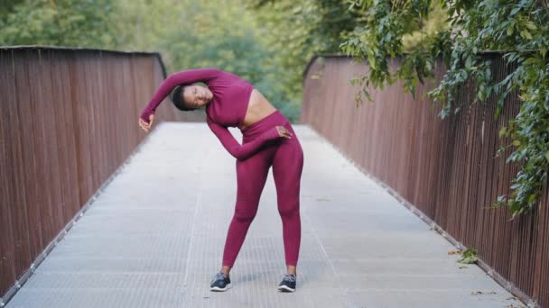 Jovem atlética em sportswear exercitando-se sozinha. Mulher adulta negra africana fazendo exercícios de alongamento durante o treino de corrida matinal no parque. Pilates, treinamento cardio, ioga e conceito de fitness — Vídeo de Stock