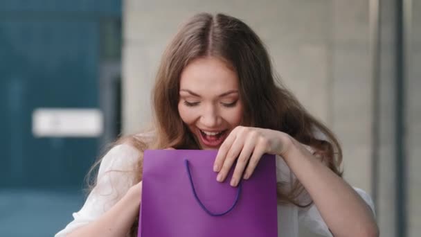 Retrato femenino al aire libre caucásico milenial joven cumpleañera dama mujer abre paquete de cartón se asoma en púrpura bolsa de regalo de compras se regocija comprando presente grita con alegría celebrando sonriente — Vídeos de Stock