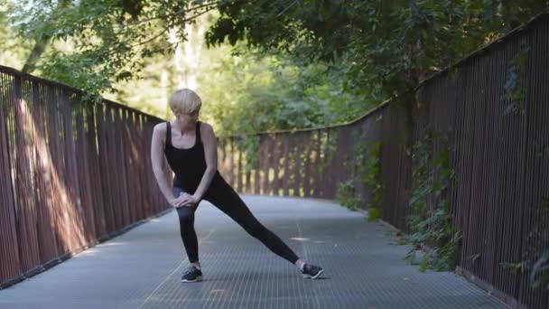 中世のブロンド白人女性アスリート女性が身に着けています黒トラックスーツスタンド市内公園で橋の上に膝のスクワットを曲げストレッチ運動を行う足の筋肉を伸ばしスポーツ屋外の柔軟性 — ストック動画