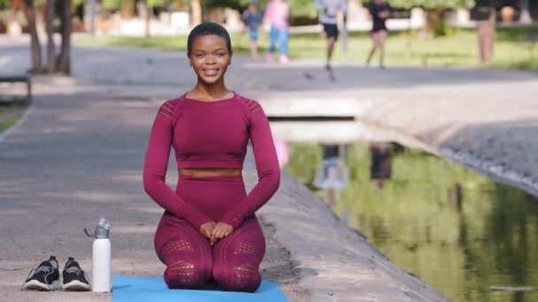 Felice donna nera sportiva che indossa abbigliamento sportivo seduto sul tappetino yoga all'aperto guardando la fotocamera, in attesa di allenamento o rilassarsi dopo l'allenamento. Plus size bellissima modella afroamericana nel parco estivo — Video Stock