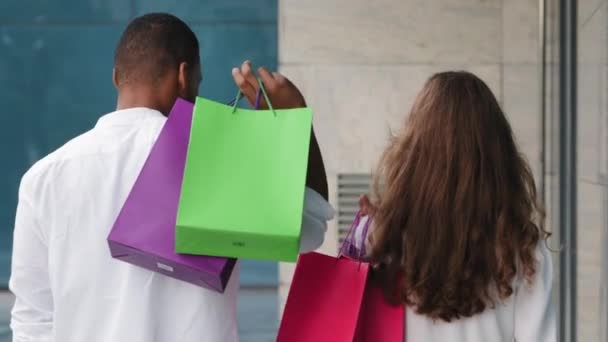 Close-up vista trasera multirracial pareja multiétnica familia africana hombre y mujer caucásica llevar bolsas de compras paquetes compra sonriendo mirando a la cámara feliz con la compra después de las ventas de la tienda de descuento — Vídeos de Stock