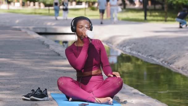 Joven atractiva mujer africana positiva más tamaño esperando entrenamiento, relajándose después de hacer ejercicio al aire libre, sosteniendo botella de agua o batido de proteínas, escuchando música con auriculares. Estilo de vida deportivo — Vídeos de Stock