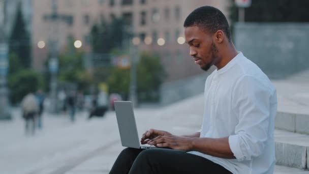 アフリカ系アメリカ人男性混合レースアフロ男民族男性学生フリーのビジネスマンの上司のリーダーライターは、市内の階段の上に座っている屋外タイプのラップトップチャットオンラインでリモートでオープンエアで作業 — ストック動画