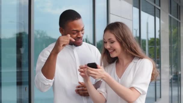 Çok ırklı çift portresi Afro-Amerikan erkek ve Kafkasyalı kadın iki arkadaş açık havada durup internetteki telefon görüntülerine bakıp gülüyorlar. — Stok video