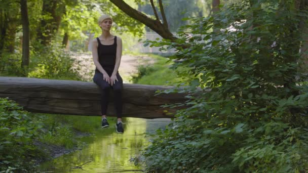 Middelbare leeftijd volwassen vrouw Kaukasische dame atleet in zwart trainingspak zitten in de zomer in het park op boomstam boven de rivier de natuur groene bomen genieten van rust frisse lucht buiten beweegt schouders dansen rusten — Stockvideo