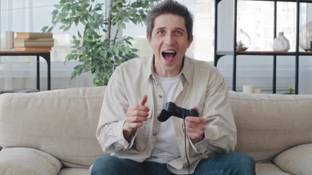 Vit man man man tusenårig kille gamer sitter hemma på soffan spela TV-spel konsol med hjälp av controller joystick njuter av spel skrik med glädje firar vinna gör seger hand gest — Stockvideo