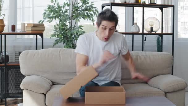 Kaukasische erwachsene männliche Käufer Kunde Millennial Mann erhält Paket von Lieferauftrag Karton Öffnen Auspacken Geschenk bekommt Angst Schreie aus Angst fühlt sich Horror versteckt hinter Sofa braucht Hilfe — Stockvideo