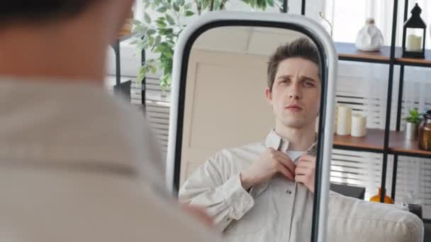 Jonge volwassene Kaukasische millennial man guy krijgen gekleed ziet er in spiegel thuis ontevreden met reflectie slechte verschijning unbutton knoppen op casual shirt voor te bereiden op datum gaan voor wandeling vergadering — Stockvideo