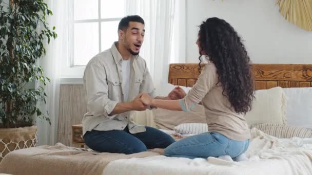 Árabe español hombre y mujer, novio y novia, marido étnico y esposa, pareja casada sentada en la cama en el acogedor dormitorio en casa cogidos de la mano sonreír hablando compartir buenas noticias disfrutar de la conversación — Vídeo de stock