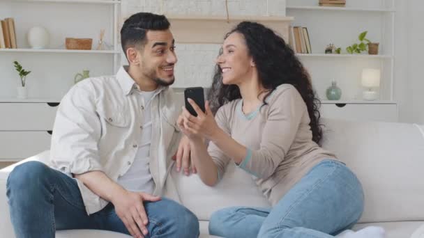 Ισπανόφωνος άνθρωπος της οικογένειας ισπανόφωνος και εθνοτική όμορφη σγουρή γυναίκα πολυφυλετικό ζευγάρι κάθεται στον καναπέ στο σπίτι κορίτσι σαλόνι κρατώντας το κινητό τηλέφωνο αναζητούν μαζί στην οθόνη smartphone μιλώντας έκπληκτος — Αρχείο Βίντεο