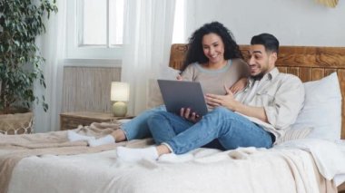 İspanyol Arap evli çift yatak odasında rahat bir yatak odasında dizüstü bilgisayarı kullanarak bilgisayar uygulamasında internet alışverişi yaparak ekranda gülümseyen bir film seçiyor.