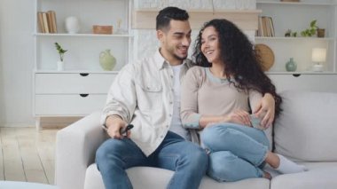 İspanyol çift evde kanepede oturmuş film seyrediyor. İspanyol koca uzaktan kumandayla ve İspanyol kadın eşinin kucaklaşması kanepede oturup gülmesi TV programını tartışması ekranda parmaklarını gösteriyor.