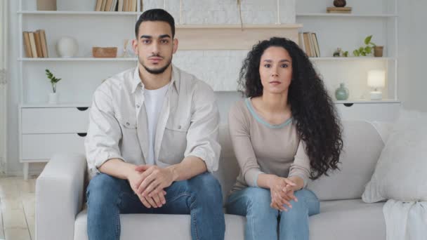 Ernsthaft fokussierte, emotionslose multiethnische Eheleute, die auf dem Sofa im heimischen Wohnzimmer sitzen, arabisch spanischer Mann und kaukasische Frau mit lockigem Haar, die entspannt auf der Couch in die Kamera schauen — Stockvideo