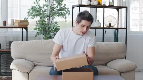 Kaukasischer lustiger Millennial-Typ Erwachsener Mann, der zu Hause sitzt, erhält Paket von Kurier öffnet Auspacken Karton gibt vor, unsichtbare Produktkleidung in Händen zu halten zeigt ok Geste Klatschen in Handflächen — Stockvideo