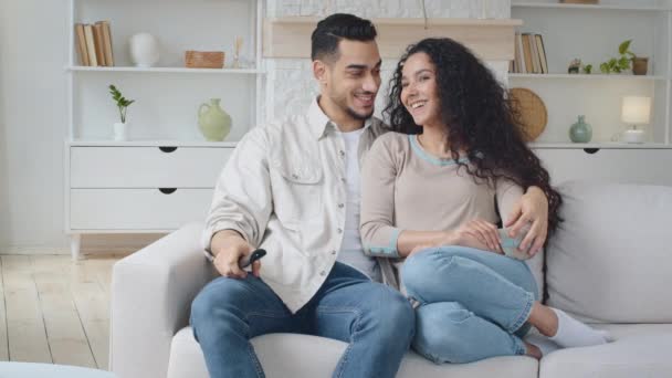 Spaans stel zit thuis op de bank en geniet van de film. Spanjaard man man man met afstandsbediening en Spaanse vrouw knuffelen zitten op de bank lachen bespreken tv-programma met vingers op het scherm — Stockvideo