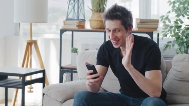 Homem branco milenar blogueiro sentado em casa sofá em quarentena lockdown fala em câmera de telefone celular faz videoconferência on-line remotamente smartphone beijos tela ondas Olá adeus — Vídeo de Stock