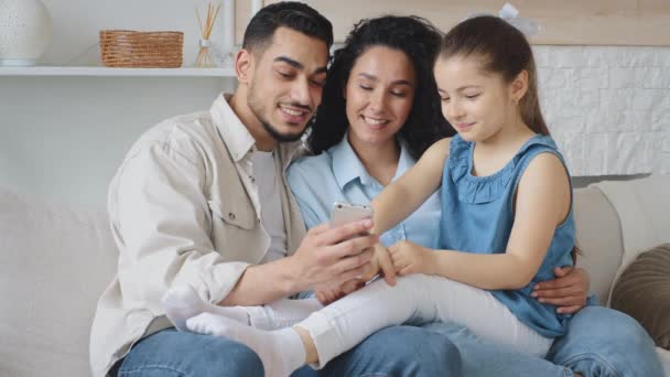 Multiracial rodina španělština španělština rodiče kavkazský dcera dítě dívka sedí na domácím gauči drží mobilní telefon mluvit s smartphone web kamera video hovor on-line konference pomocí aplikace — Stock video
