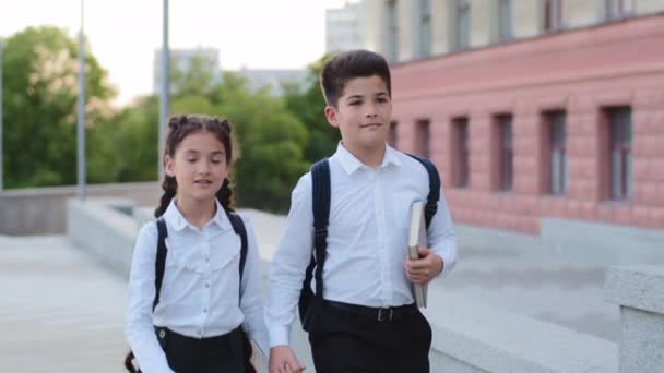 Two children schoolchildren hispanic pupils kids little girl and boy with book brother and sister friends wear school backpacks holding hands walking together down street outdoors to lesson for class — Stock Video