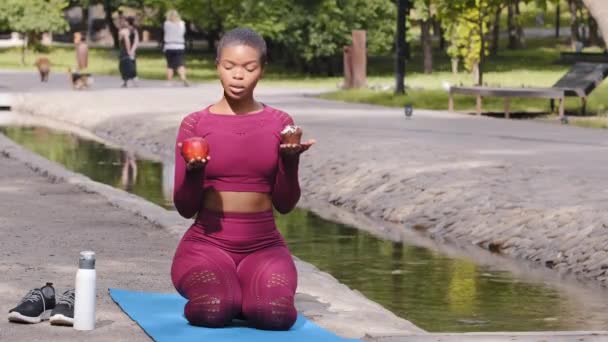 Plus size model Afro-Amerikaanse mooie vrouw zittend op yoga mat na training in zomerpark met keuze tussen appel en cupcake. jong afro meisje aarzelend over keuze van taart of fruit — Stockvideo