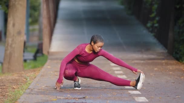 Portret afrykańskiej sportowej kobiety wykonującej ćwiczenia rozciągające w parku przed treningiem. Sportowiec przygotowujący się do joggingu na świeżym powietrzu. Biegacz rzuca się z boku. Koncepcja aktywnego stylu życia sportu — Wideo stockowe