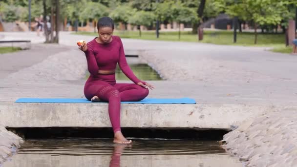 Menina africana atlética confiante millennial em sportswear relaxante no tapete de ioga após o treino. Instrutor de fitness atraente jovem mulher adulta relaxa, come maçã, escolhe comida saudável, estilo de vida ativo — Vídeo de Stock