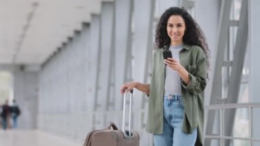 Güzel İspanyol kadın cep telefonunda kısa mesajlar yazarak havaalanı terminalinde cep telefonu ve bavul taşıyan bayan turist saatlerini kontrol ediyor.
