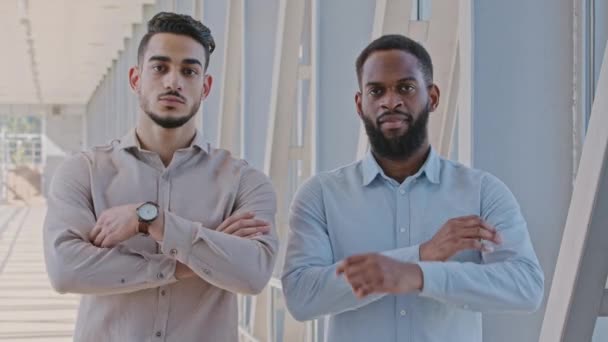 Két multiracionális férfi barát kollégák hispán férfi és afro-amerikai szakállas fickó formális ingben áll zárt ajtók mögött, komoly arckifejezéssel keresztbe tett karokkal nézi a kamerát. — Stock videók