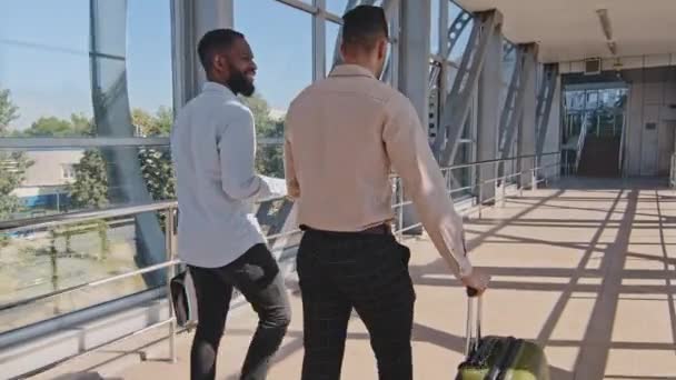 Dos hombres amigos multirraciales colegas hombres socios de negocios hombre hispano inversionista llevando equipaje maleta y afroamericano chico líder vistiendo camisas formales caminando en terminal del aeropuerto hablando — Vídeo de stock